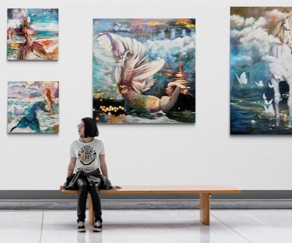 woman stting in gallery on bench surrounded by paintings on wall