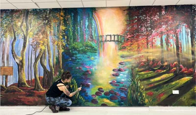 Woman Working on mural of forest stream with bridge and trees with pink and red leaves