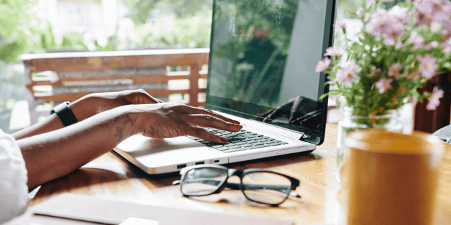 hands typing on computer with glasses on wood table pink flowers in vase