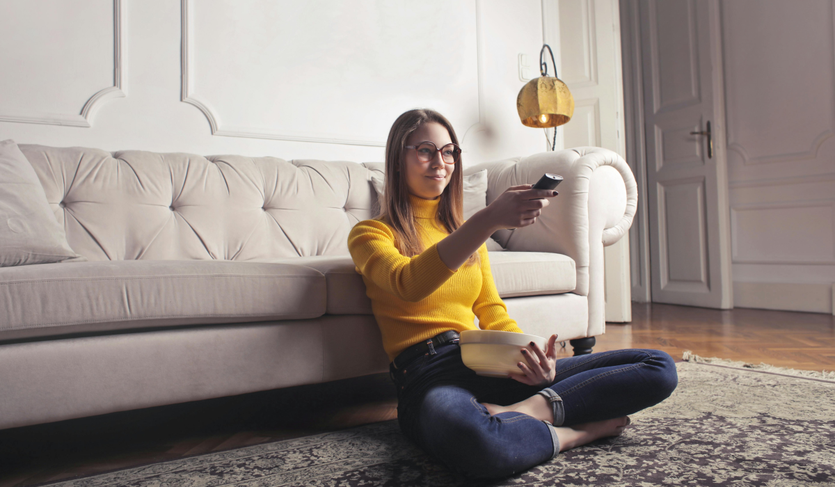 Woman sitting in yellow swaeter pointing tv remote at tv sitting up against a cream couch 