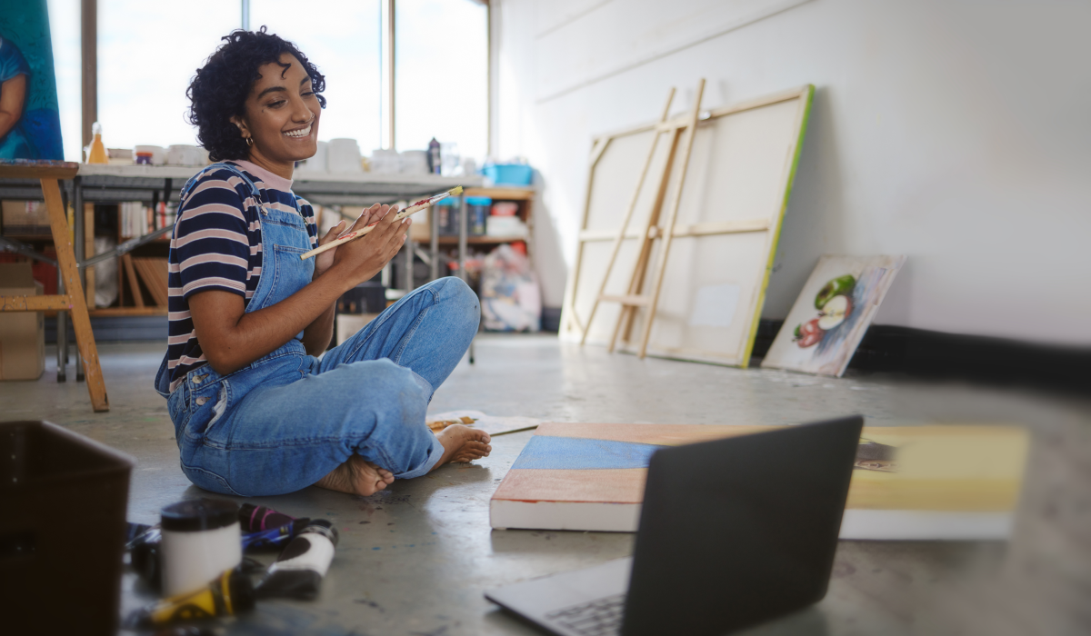 Woman sitting in front of painitng canvases in blue overalls 