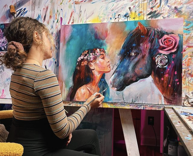 girls in tan shirt painting sitting in frint of painting of a brown horse and girl with flower crwon on 