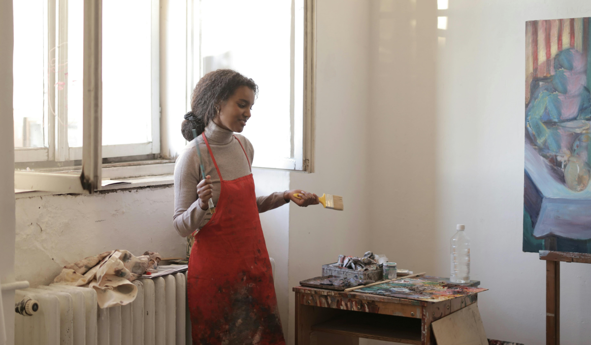 WOman in painting apron standing in front of window with paing on pallet and holding paint brush